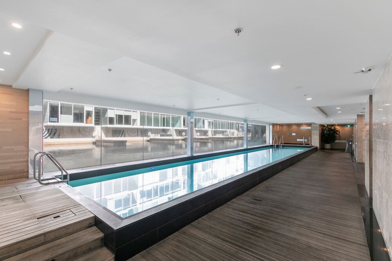 En modern inomhuspool med väggar av genomskinligt glas, trägolv och utsikt över poolområdet.