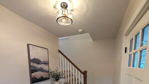 A hallway with a wooden floor, a staircase with white railings, a wall-mounted light fixture, and a door with a window.