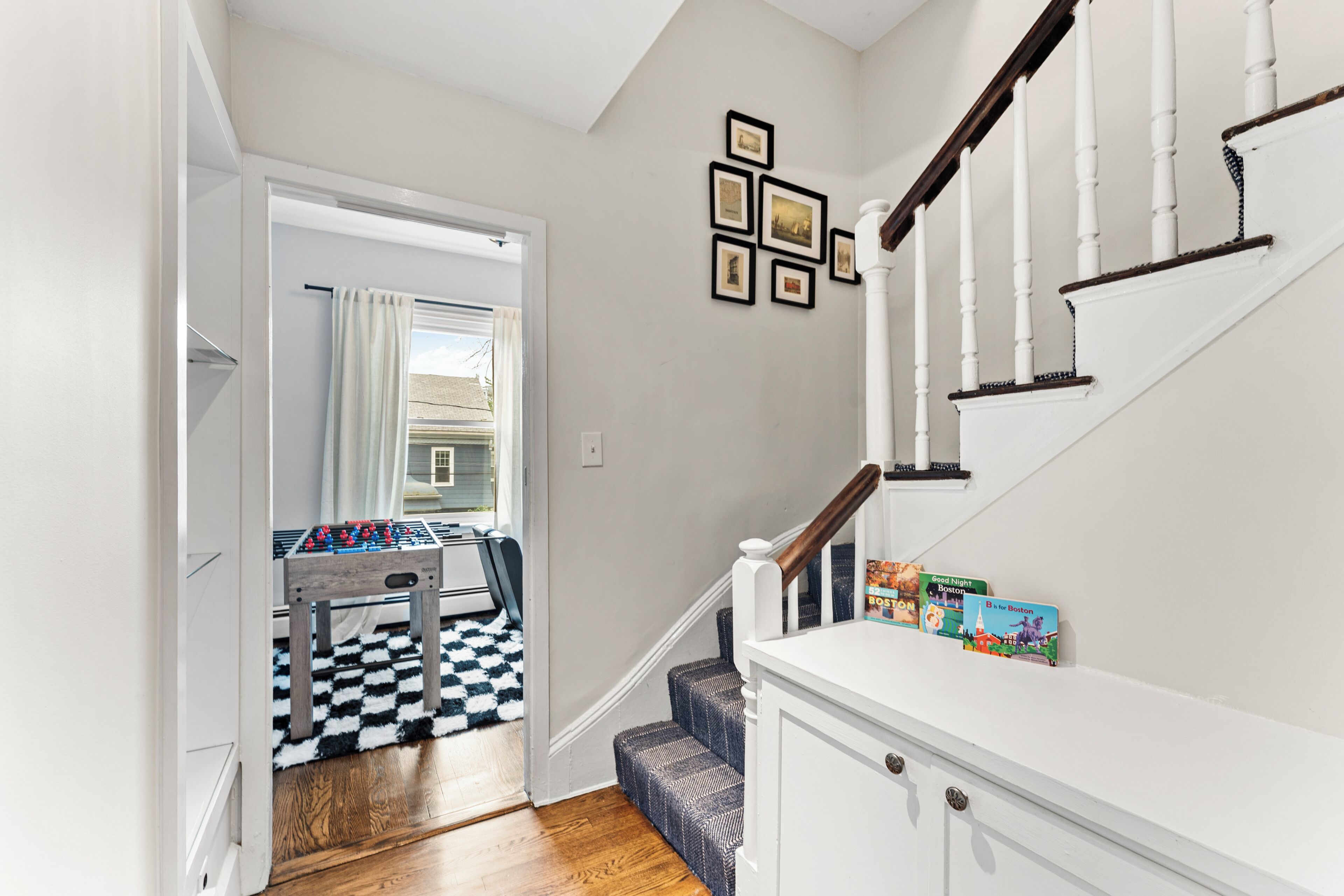 A hallway with a staircase, a checkered rug, and a set of framed pictures on the wall.