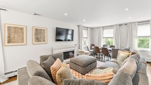 Una sala de estar moderna con un sofá gris, un otoman y almohadones decorativos, un televisor de pantalla plana, una chimenea y un comedor con sillas y una mesa.