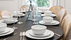 Una mesa de comedor preparada con platos, tazones y copas de vino, rodeada de sillas.