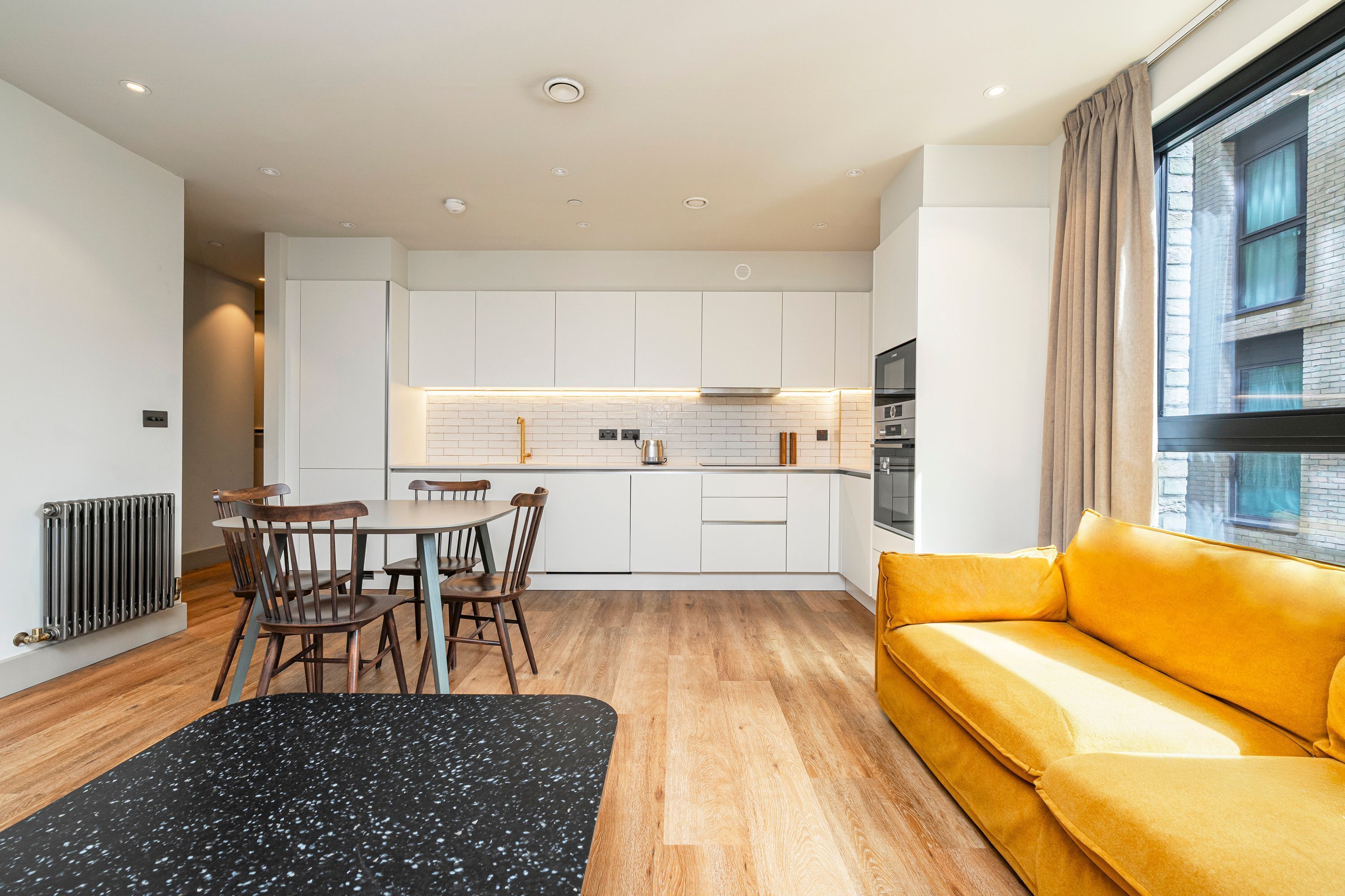 A modern kitchen with white cabinets, a dining table with chairs, a yellow sofa, and a black rug.