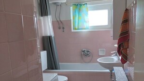 Et lille badeværelse med vaskemaskine, toilet, badekar og et vindue med gardin.