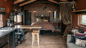 Un dormitorio acogedor con techo de madera, una cama con muchos almohadones, un taburete pequeño, una mesita de noche con lámpara y una ventana con cortinas.