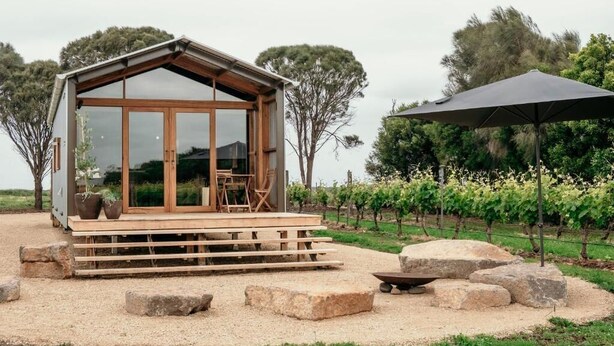 Una cabaña moderna con amplios ventanales, una terraza de madera y un viñedo al fondo.