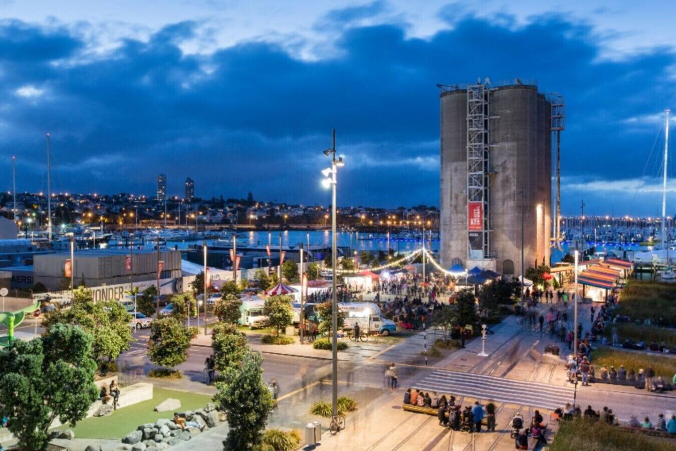 A waterfront urban area with a large concrete structure, a marina, and a public square with trees and seating.
