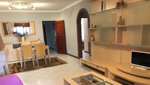 A living room with a TV, a glass coffee table, a dining area with chairs, and a kitchen in the background. - 2-bedroom Quarteira apartment near the beach. (Quarteira)