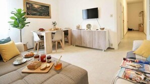 Una sala de estar con un sofá, una mesa de comedor con sillas, un televisor montado en la pared y un pasillo que da a otra habitación.