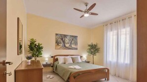 Een slaapkamer met een bed, een plafondventilator, een schilderij, een potplant en een raam met gordijnen.