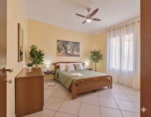 Ein Schlafzimmer mit einem Bett, einem Deckenventilator, einem Gemälde, einer Topfpflanze und einem Fenster mit Vorhängen.