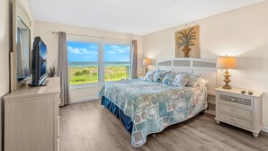 A bedroom with a bed, a TV, a nightstand, and a window with a view of the ocean. - Sunrise Oceanfront Condo 3BR | Balcony Breezes and Heated Pool 🌞🍹 (Cocoa Beach)