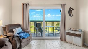 A room with a sliding glass door to a balcony, a reclining chair, a small table, and a cabinet. - Sunrise Oceanfront Condo 3BR | Balcony Breezes and Heated Pool 🌞🍹 (Cocoa Beach)