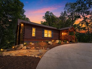 A log cabin with a porch, stone pathway, and a view of the surrounding forest.