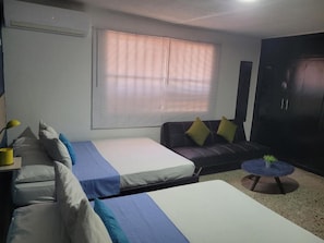 Una habitación con dos camas, un sofá, una mesa redonda con una planta y una ventana con persianas.