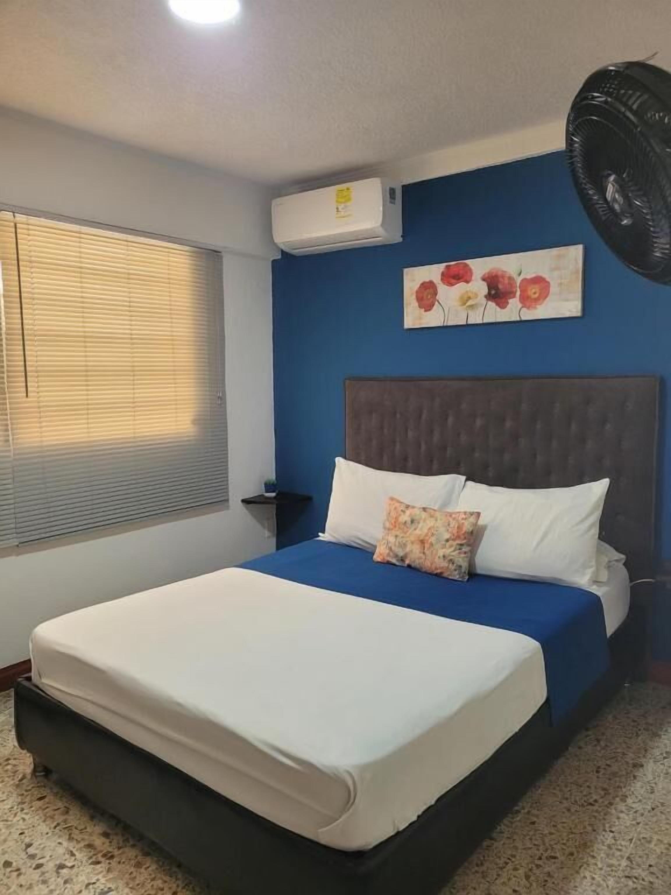 Un dormitorio con cama, aire acondicionado de pared, ventilador y ventana con persianas.