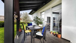 Eine Terrasse im Freien mit Tisch und Stühlen, einem überdachten Bereich und einer Glastür zum Innenbereich.