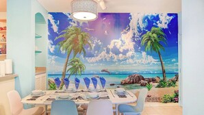A dining area with a mural of a beach scene, a chandelier, and a table set for four. - Bright 3BR Resort Condo Near Disney with Pool, Gym, Balcony (Kissimmee)