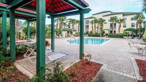 A covered outdoor area with a swimming pool, lounge chairs, and a building with multiple stories. - Bright 3BR Resort Condo Near Disney with Pool, Gym, Balcony (Kissimmee)