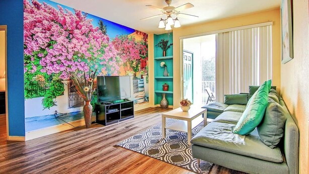 A living room with a television, a sofa, a coffee table, a patterned rug, and a wall mural. - Bright 3BR Resort Condo Near Disney with Pool, Gym, Balcony (Kissimmee)