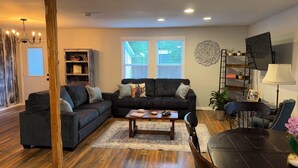 A living room with a dark grey sofa, a wooden coffee table, and a dining table with chairs. There is a bookshelf, a ladder, and a decorative wall piece.