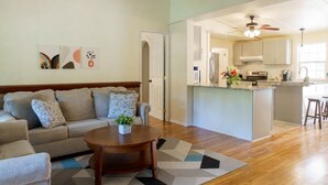 Una sala de estar con sofá, una mesa de centro y una alfombra. Incluye una zona de cocina con refrigerador, estufa y taburetes de bar.