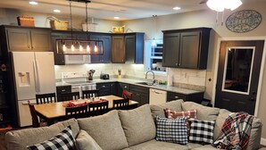A modern living room with a sectional sofa, a small wooden table, and a kitchen area with dark cabinets and a white refrigerator. - Bunk/Game Room, Near Lake, Pet Friendly, Firepit, FUN! (Toccoa)