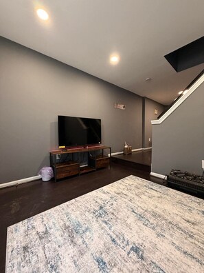 Een woonkamer met een flatscreen televisie op een houten standaard, een kamerbreed tapijt met een patroon en een trap met een zwarre leuning.