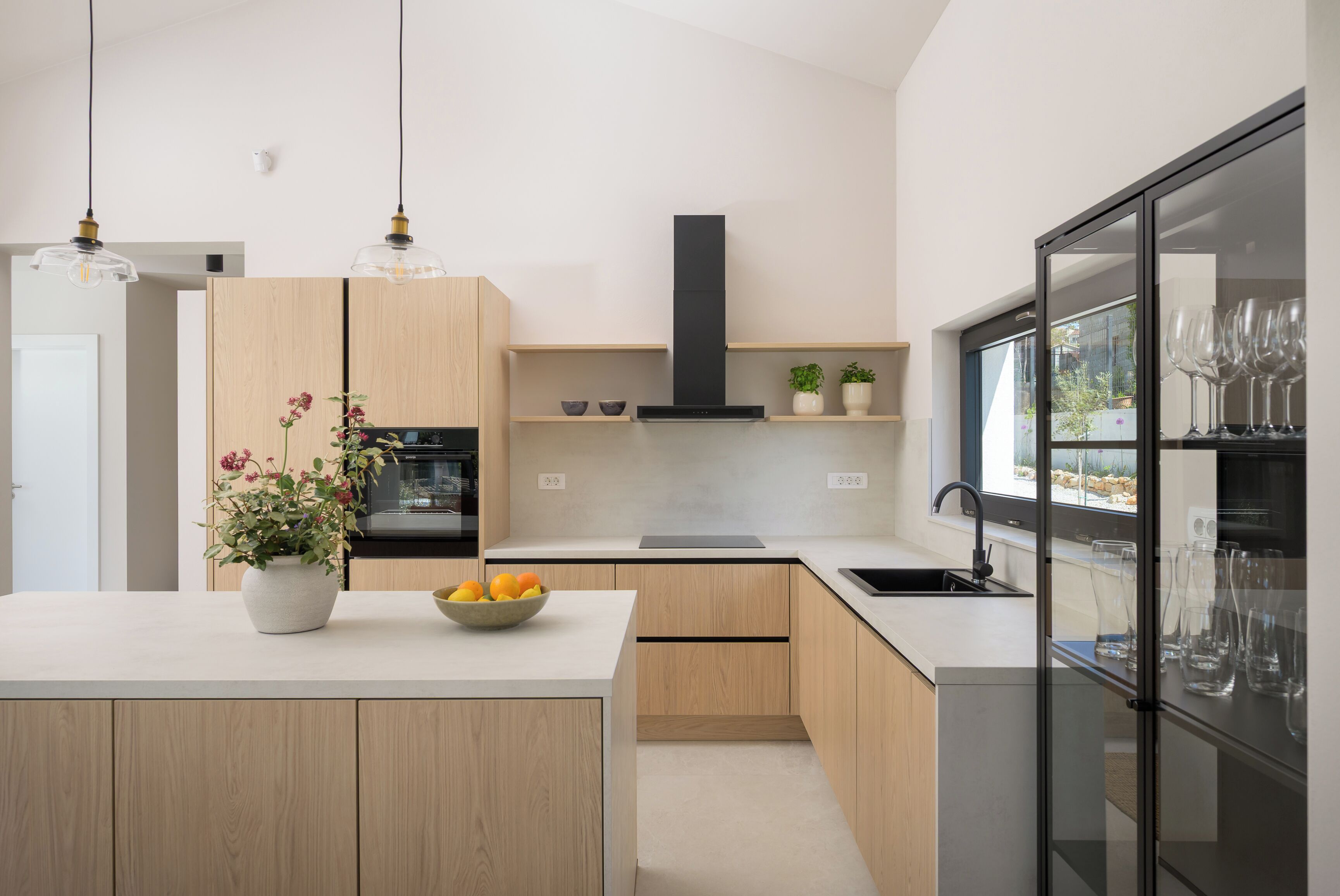 Une cuisine moderne avec des meubles en bois, un îlot central, une hotte noire et un meuble à vin avec une porte en verre.