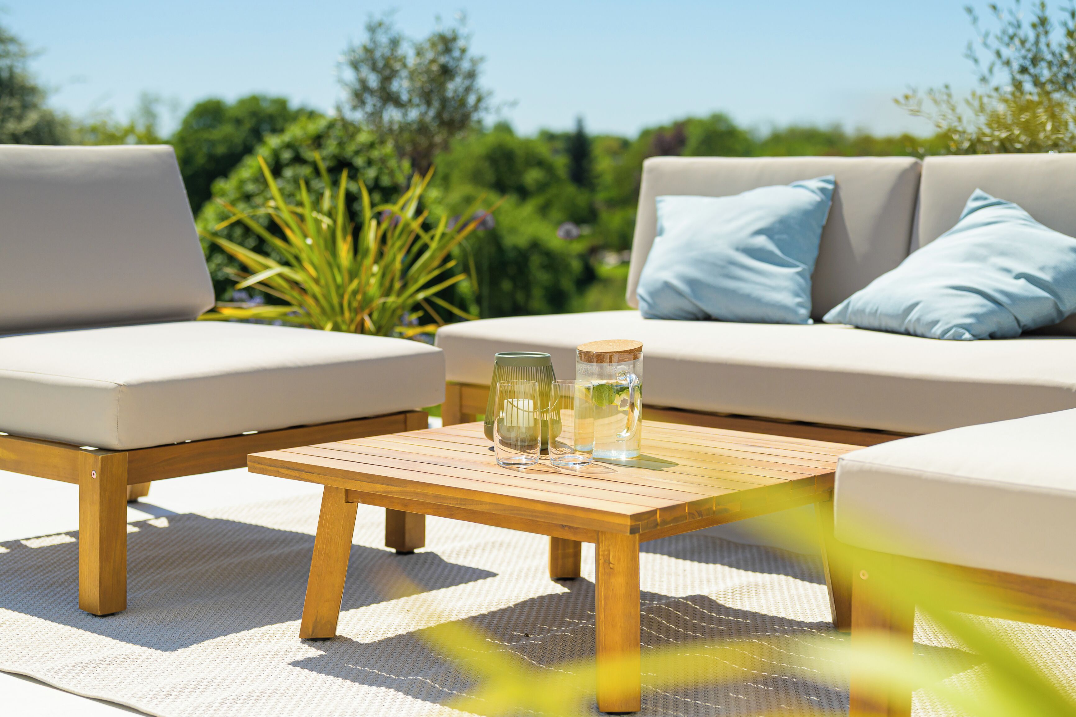 A sunlit outdoor seating area with a wooden table, two cushioned chairs, and a sofa with light blue cushions.