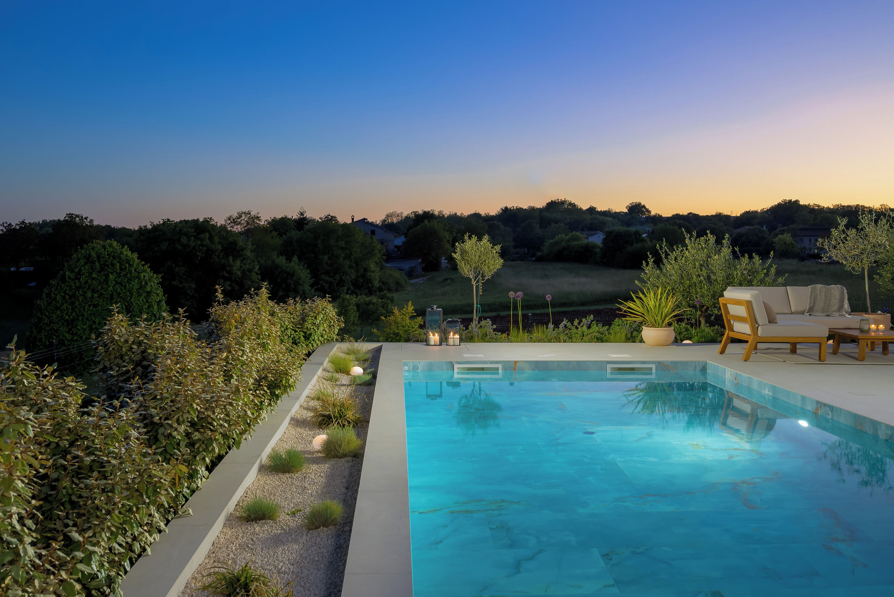 Una piscina rettangolare con superficie d'acqua cristallina, circondata da un basso siepe e un cordolo in ghiaia. Due lettini prendisole e un tavolino con una lanterna sono a bordo piscina.