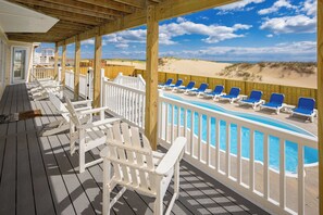 Eine Holzterrasse mit weißen Geländern, ein Pool mit blauen Liegestühlen und Blick auf den Strand.