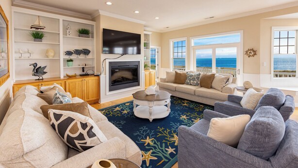 A living room with a fireplace, sofas, a coffee table, and a view of the ocean. - Dmanfish - Oceanfront 9 BR w/pool (Virginia Beach)