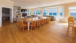 A spacious dining area with a large wooden table and chairs, a kitchen with a TV, and a view of the sea through large windows. - Dmanfish - Oceanfront 9 BR w/pool (Virginia Beach)