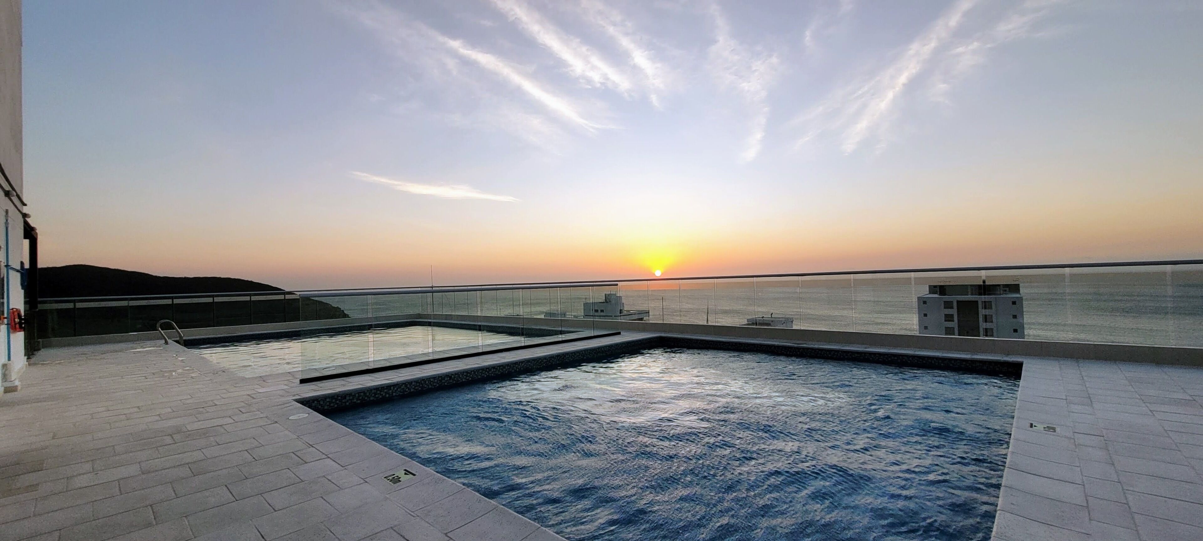 A rooftop pool with a sunset view, a glass railing, and a small building structure.