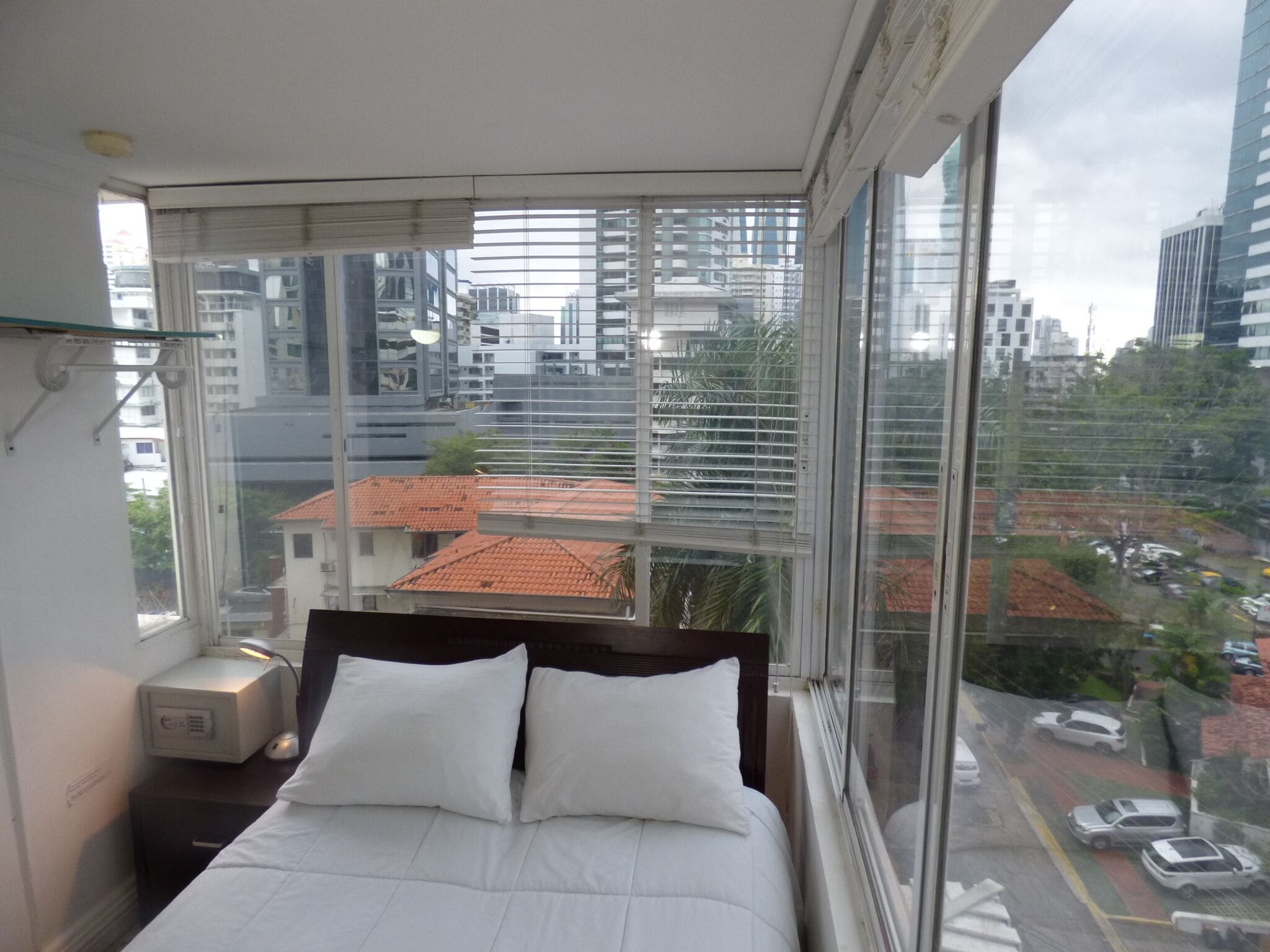 A bedroom with a large window offering a city view, a bed with white bedding, and a bedside table with a lamp.