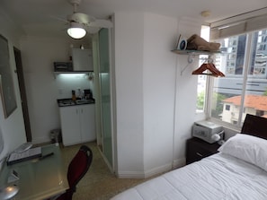 Ein kleines Hotelzimmer mit Küchenzeile, einem Bett, einem Schreibtisch und einem Fenster mit Blick auf Gebäude.