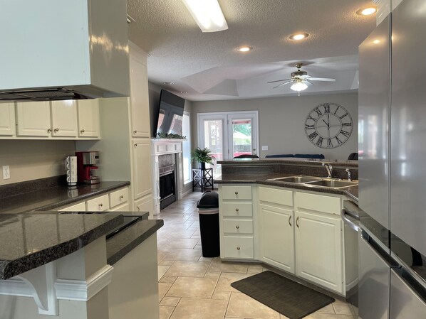 Una cocina con armarios blancos, encimera de granito, refrigerador de acero inoxidable, ventilador de techo y un reloj de pared.