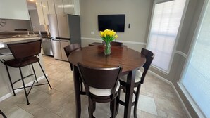 A modern kitchen with a dining area, including a round wooden table with chairs, a refrigerator, and a flat-screen TV mounted on the wall. - Beautiful 3-bedroom home near Southeast hospital and downtown Dothan (Dothan)