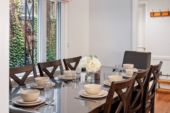 A dining table set for six with white plates, bowls, and glasses, surrounded by wooden chairs.