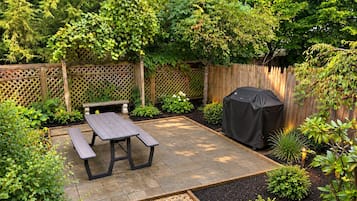 Un patio con una mesa de picnic y una parrilla, rodeado por una valla de madera y diversas plantas.