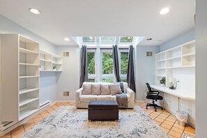 A modern living room with a sofa, ottoman, and a desk with a chair. There are built-in shelves on the walls and a large window with curtains.