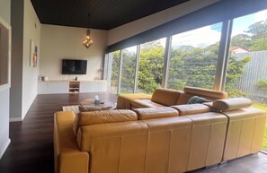 A modern living room with a leather sofa, a flat-screen TV, and a dining area with a wooden table. - Jasmine Escape |Architect-Designed Bush Retreat, Pool, Sunken Spa & Fire Pit (Ellalong)