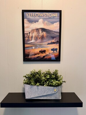 Een lijst met een poster van Yellowstone National Park aan de muur, een grijze vilten plantenbak met groene planten eronder, en een zwart schap.