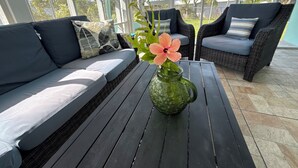Un patio iluminado por el sol con un sofá de mimbre, una mesa de vidrio y un florero con una flor.