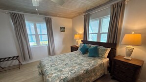 Un dormitorio con una cama, dos mesitas de noche, una lámpara de pared y un ventilador de techo, además de un ventanal con cortinas.