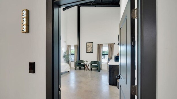 An open door to a room with a bed, two chairs, a table, and a window with curtains.