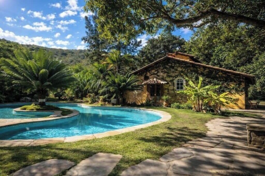 A pool with a curved edge, a stone pathway, and a thatched-roof building surrounded by lush greenery.