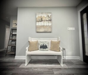 Um banco branco com duas almofadas, um quadro fixado na parede e uma escada de metal em um corredor.