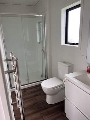 Un baño con cabina de ducha de vidrio, inodoro blanco y lavamanos con ventana superior.