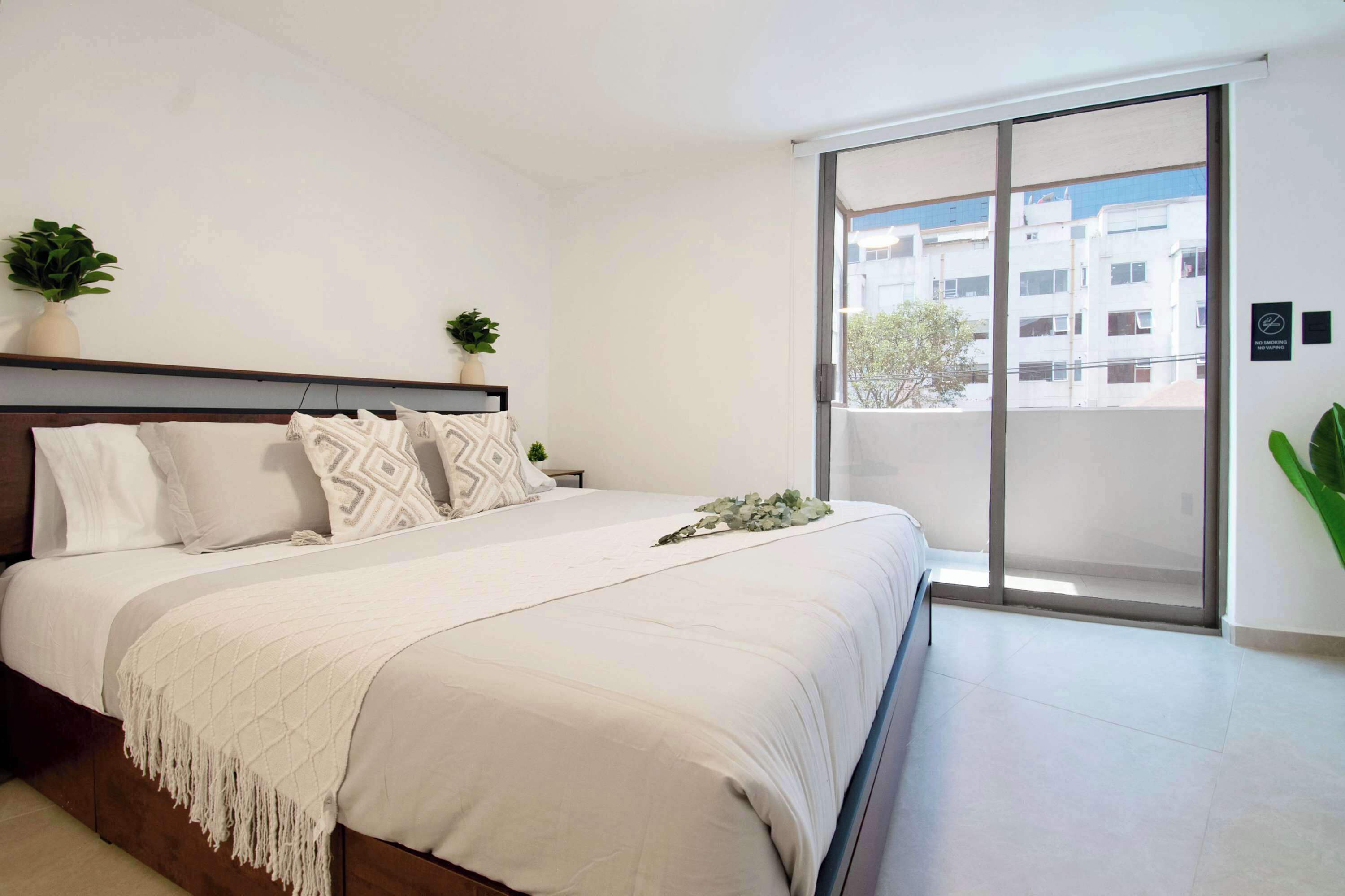 A bedroom with a bed, a nightstand, a potted plant, a sliding glass door, and a view of buildings outside.